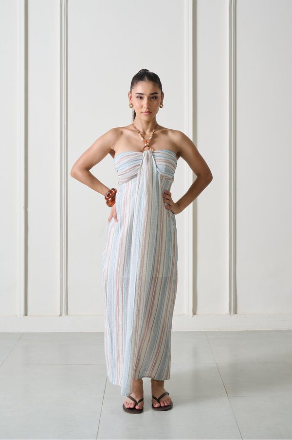 A woman modeling a strapless maxi dress with vertical pastel stripes in light blue, pink, and beige, standing against a white paneled wall.