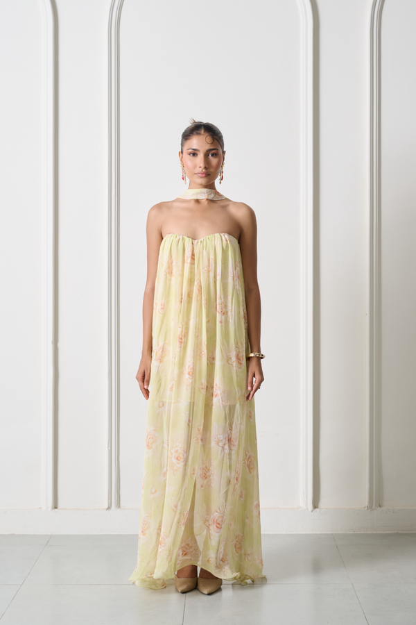 A woman stands in a white arched room wearing a strapless pale yellow maxi dress with a pink floral print and a matching choker.