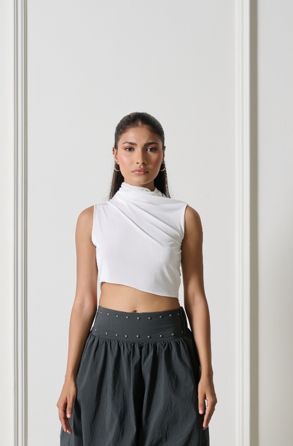 A woman modeling a white asymmetrical sleeveless crop top paired with dark gray wide-leg pants, standing against a white paneled wall.