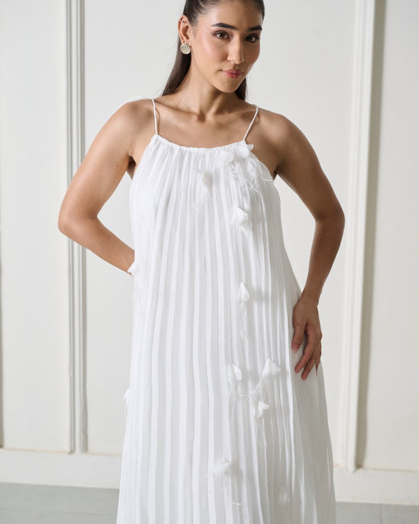 A woman modeling a white sleeveless maxi dress with thin spaghetti straps, a gathered pleated bodice, and small tassel accents, set against a light cream paneled wall.