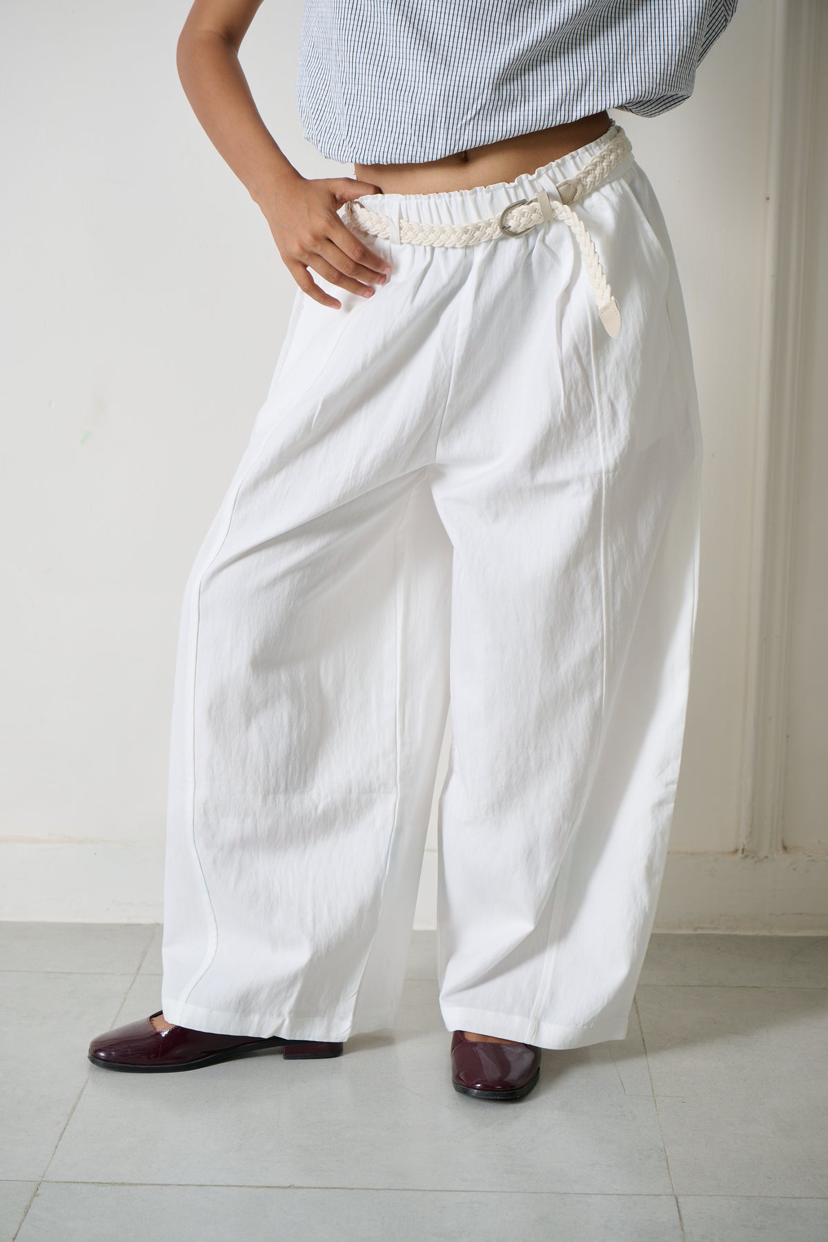 White wide-leg linen pants with an elastic waistband and a decorative cream drawstring, shown on a model wearing a light blue striped crop top and dark brown shoes.