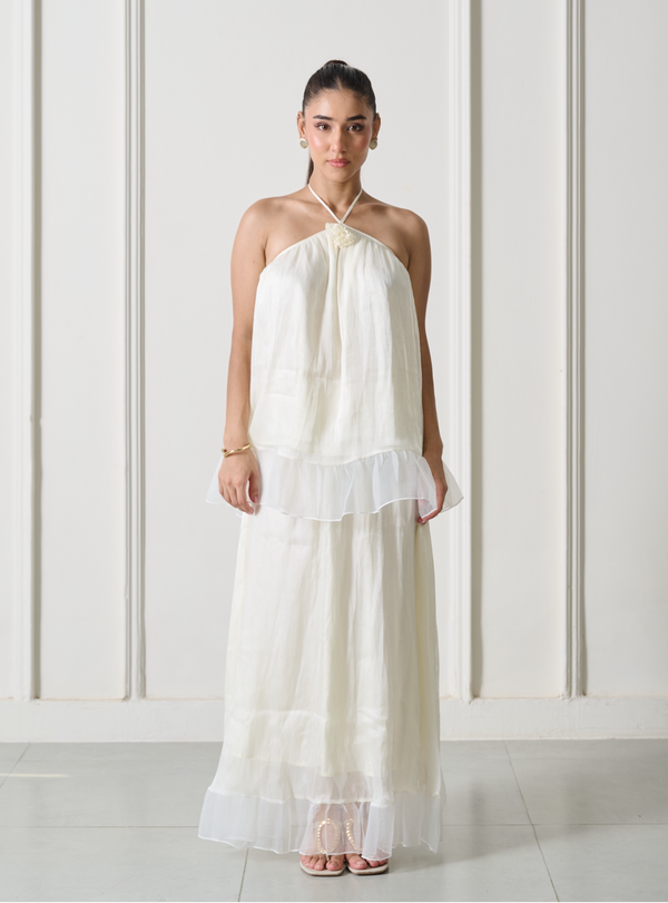 A woman modeling a white halter-neck maxi dress with a sheer ruffled overlay and tiered skirt, standing against a white paneled wall.