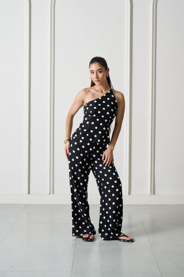 A woman modeling a black one-shoulder jumpsuit with white polka dots, standing in front of a white wall with arched recesses.
