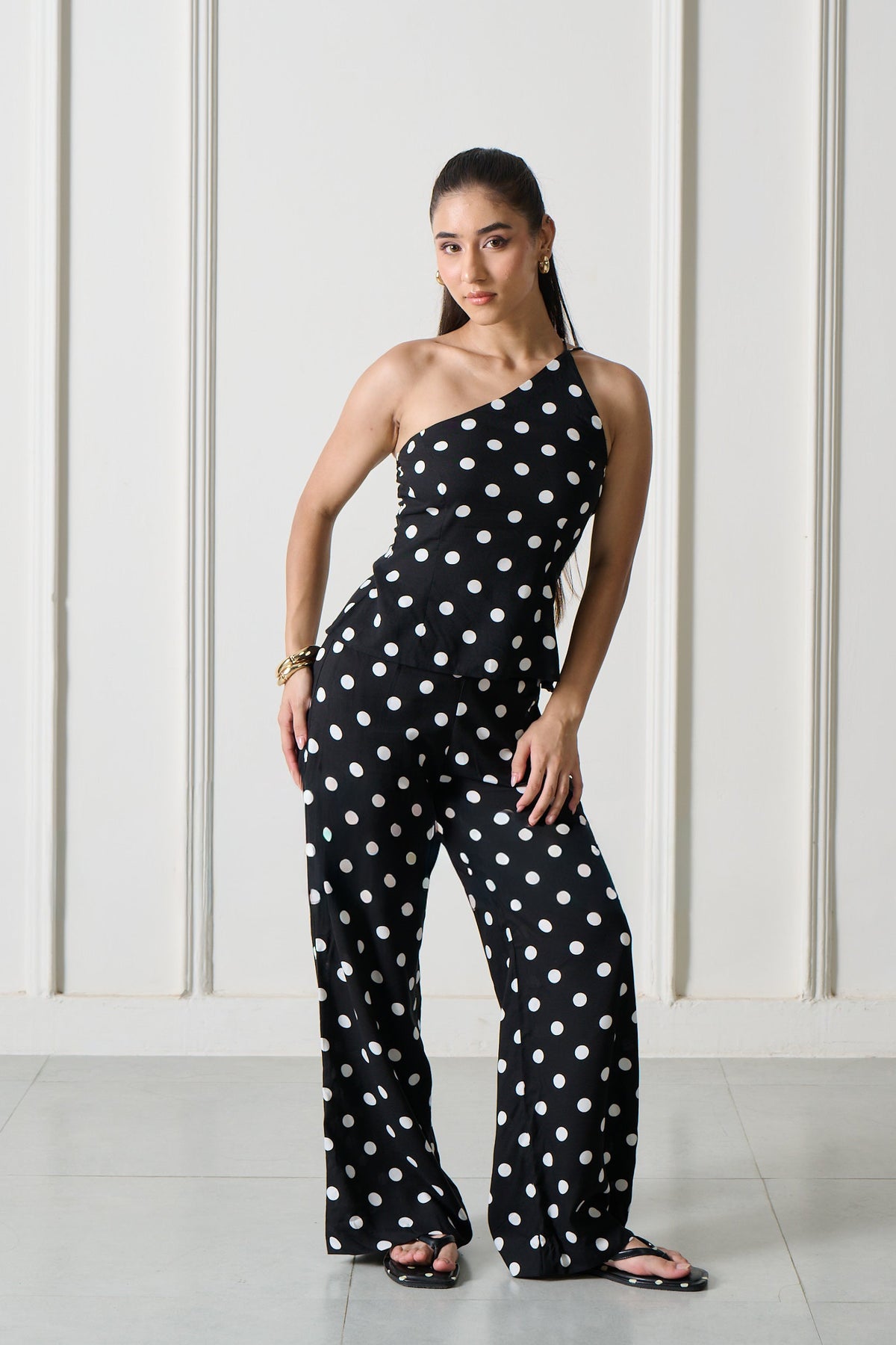 A woman modeling a black one-shoulder jumpsuit with white polka dots, standing in front of a white wall with arched recesses.