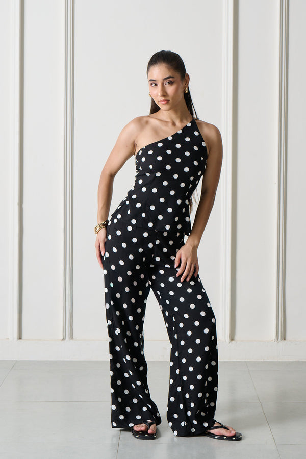 A woman modeling a black one-shoulder jumpsuit with white polka dots, standing in front of a white wall with arched recesses.