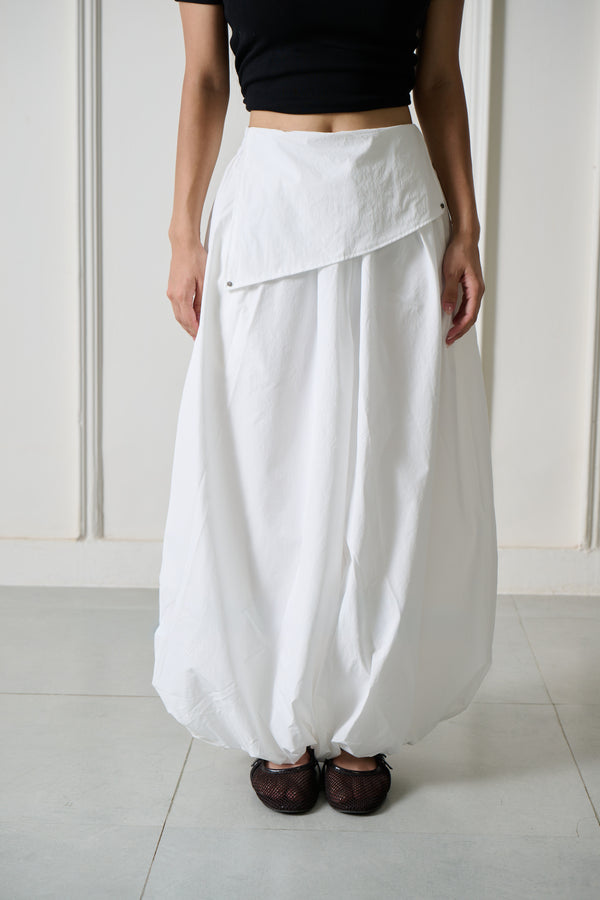A person wearing a loose, high-waisted white pant with an asymmetrical wrap-style front, paired with a black cropped top and dark brown shoes, standing against a light paneled wall.