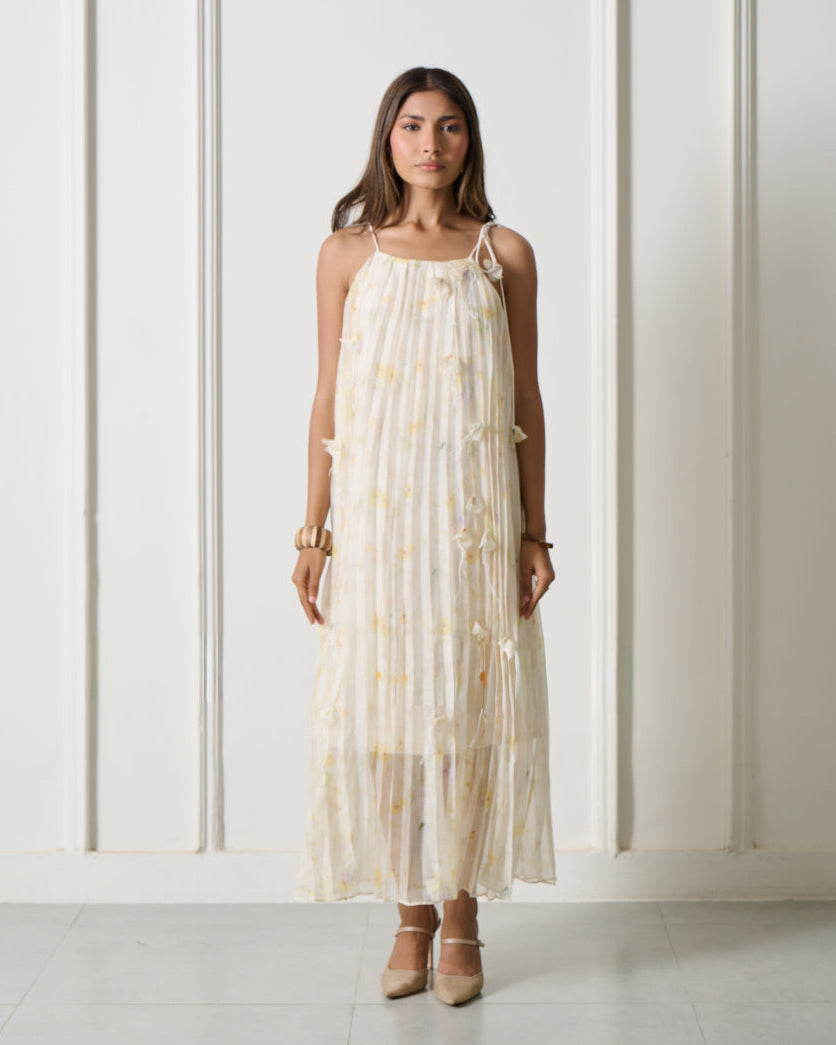 A woman stands in a bright white room with arched wall details, wearing a sleeveless cream maxi dress with a subtle yellow floral pattern and beige heels.