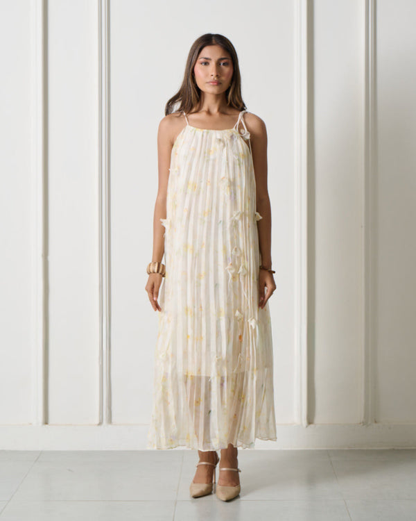 A woman stands in a bright white room with arched wall details, wearing a sleeveless cream maxi dress with a subtle yellow floral pattern and beige heels.