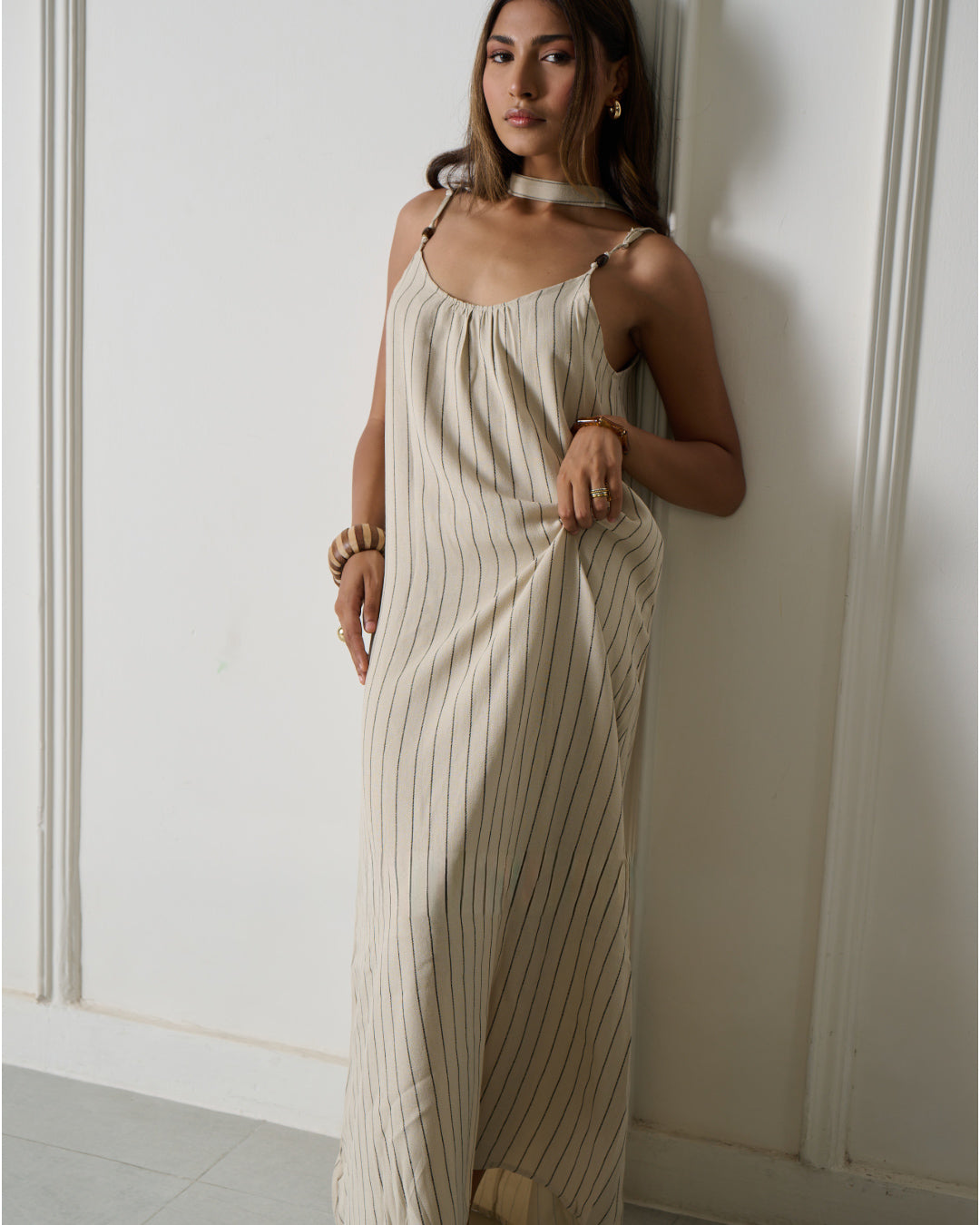 A woman wearing a cream-colored maxi dress with thin vertical stripes, leaning against a white paneled wall.