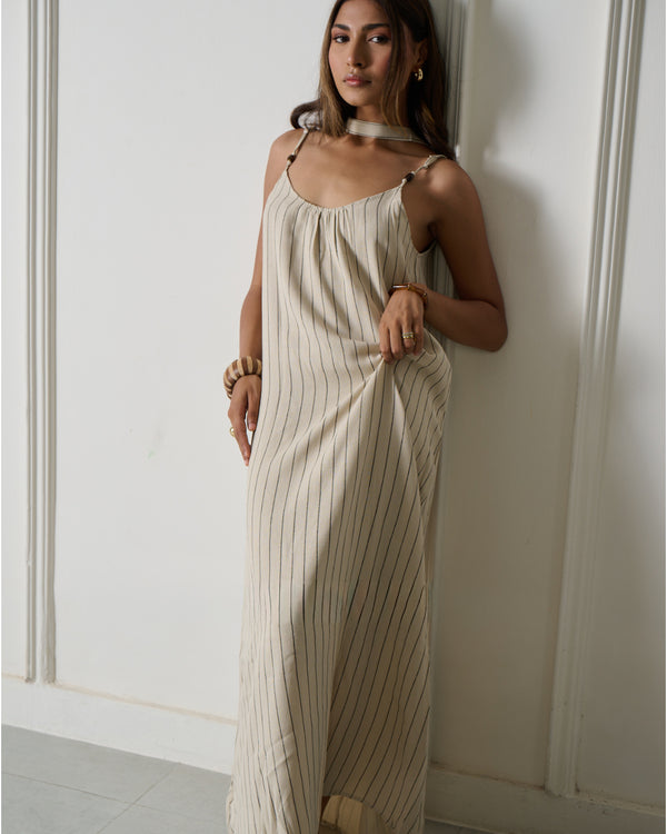 A woman wearing a cream-colored maxi dress with thin vertical stripes, leaning against a white paneled wall.