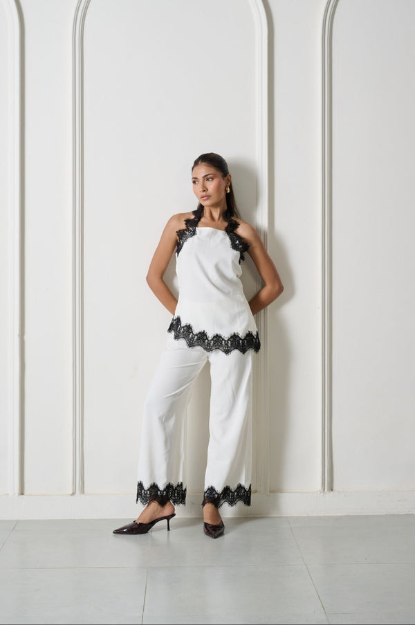 A woman stands against a white wall with three arched recesses, wearing a white sleeveless halter-style top and matching wide-leg pants with black lace trim at the halter, hem, and cuffs, paired with black pointed heels.