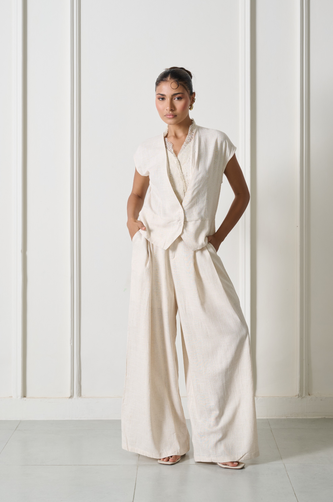 A woman modeling a cream-colored linen jumpsuit with a wrap-style vest and wide-leg trousers, standing against a white paneled wall.