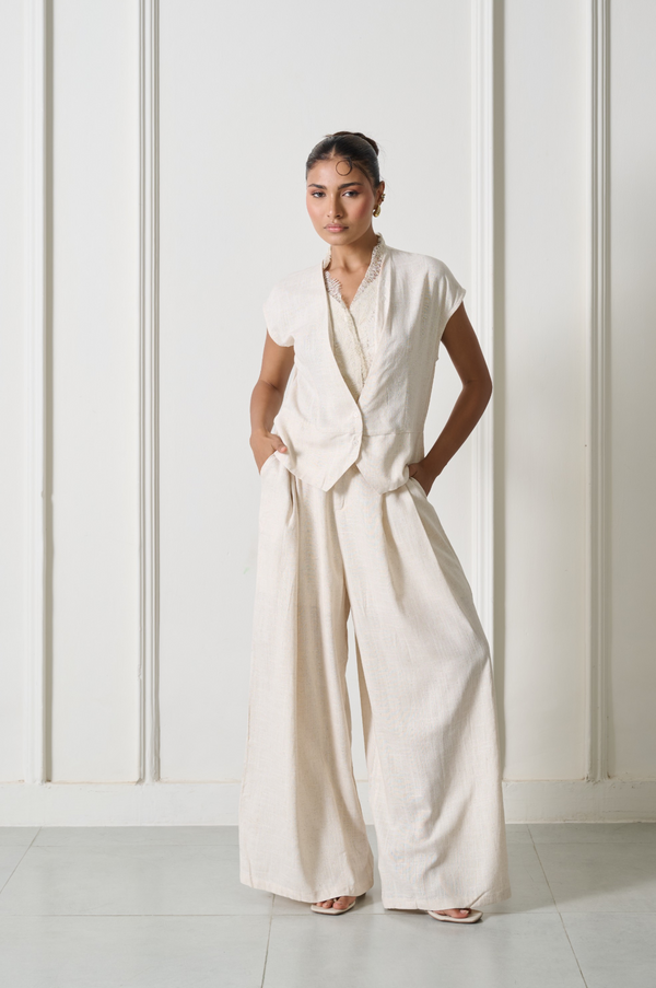 A woman modeling a cream-colored linen jumpsuit with a wrap-style vest and wide-leg trousers, standing against a white paneled wall.