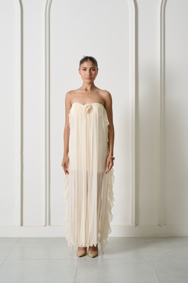 A woman stands in a studio wearing a strapless cream-colored maxi dress with a ruffled tiered skirt and a bow detail at the bust. The background is a clean white wall with vertical molding and light gray tiled flooring.