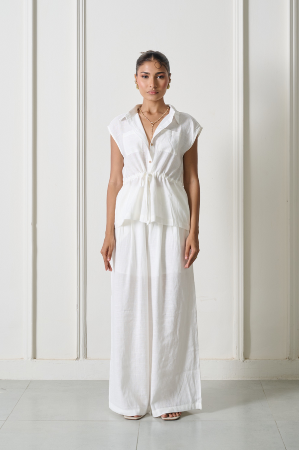 A woman stands facing forward in a white sleeveless collared top with a gathered waist and matching wide-leg white pants, photographed against a light cream paneled wall.