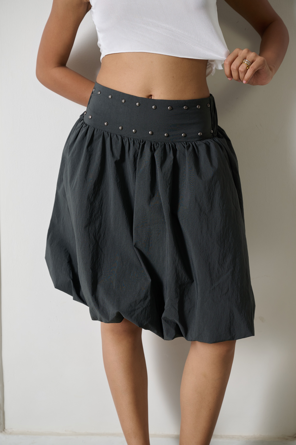 A person wearing a dark charcoal gray balloon-style skirt with a studded waistband, paired with a white cropped top, standing against a plain off-white wall.