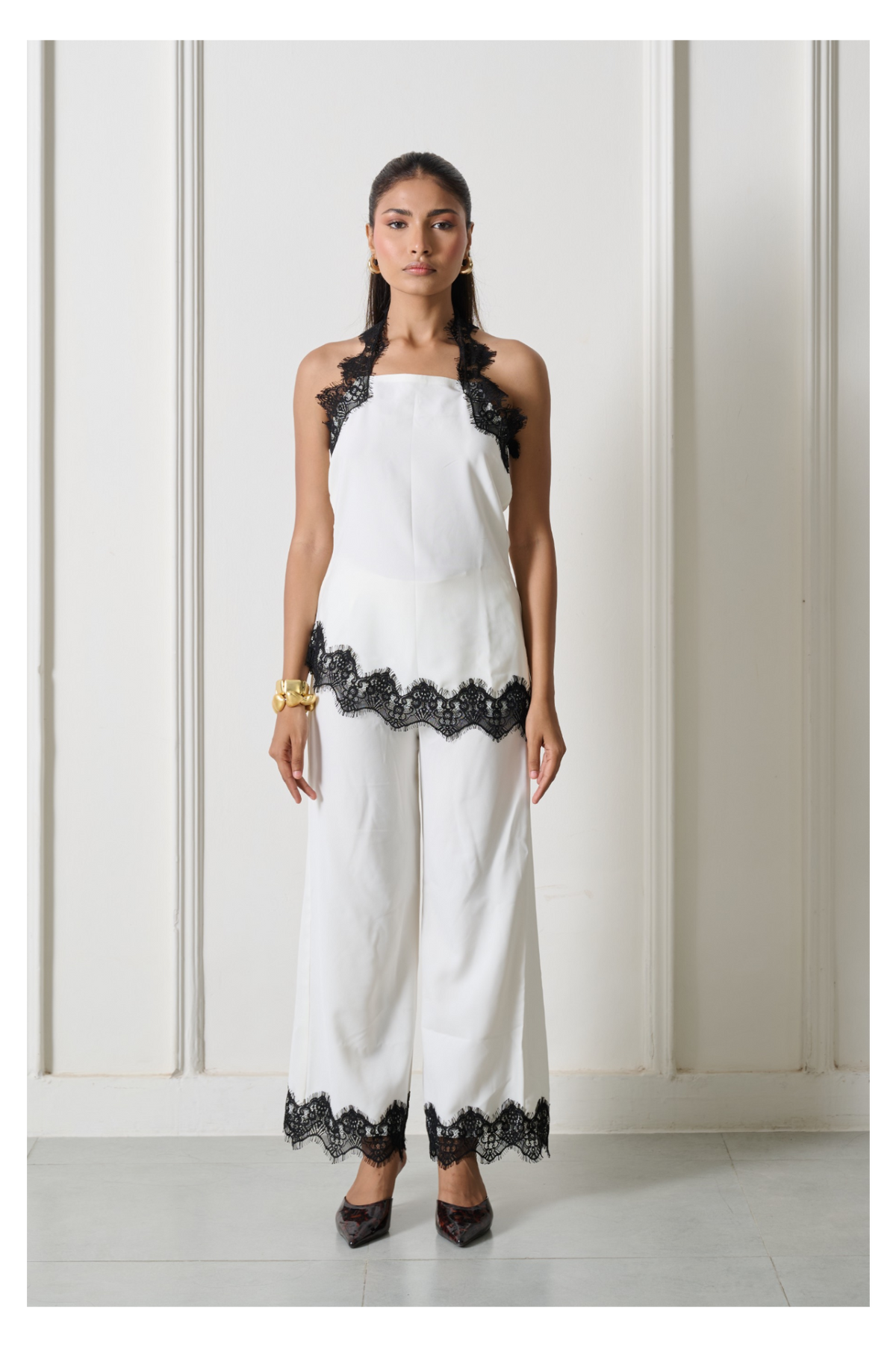 A woman modeling a white sleeveless halter jumpsuit with black lace trim at the shoulders, hem, and pant cuffs, standing against a white paneled wall.