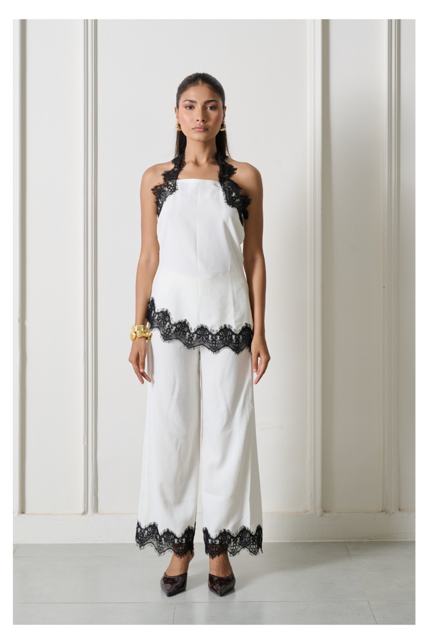 A woman modeling a white sleeveless halter jumpsuit with black lace trim at the shoulders, hem, and pant cuffs, standing against a white paneled wall.
