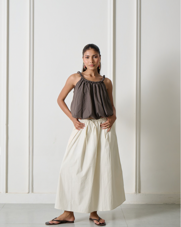 A woman modeling a dark brown sleeveless top with ruffled shoulder straps and a cream-colored maxi skirt, standing against a white paneled wall.
